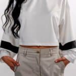 A woman wearing a stylish white long-sleeve crop top with black stripe details on the sleeves, paired with beige high-waisted pants, showcasing casual fashion.